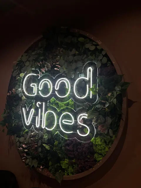 Baltas LED neoninis užrašas „Good vibes“ ant samanų sienos apskritime