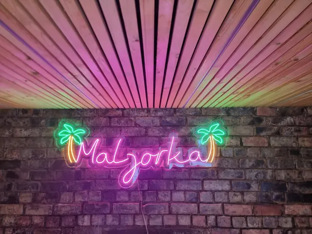 Rožinis LED neoninis užrašas su žaliomis palmėmis ant plytų sienos
